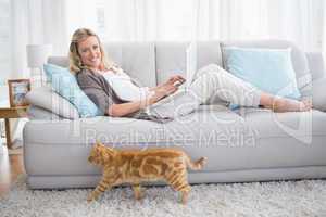 Woman lying on sofa using her laptop smiling at camera