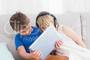 Siblings using a headphone and a tablet sitting on a couch