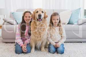 Sisters sitting on rug with golden retriever smiling at camera