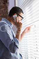 Businessman using mobile phone while looking through blinds in o