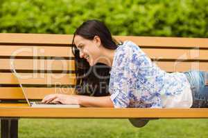 Happy brunette lying on bench using laptop