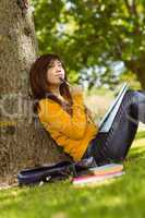 Female student with books sitting against tree in park