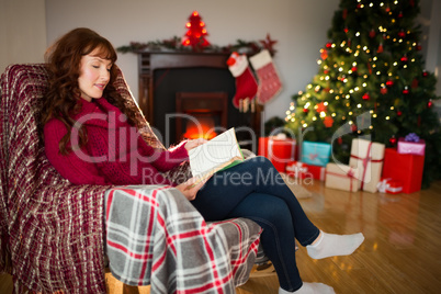 Smiling redhead reading on the armchair at christmas