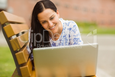 Smiling brunette lying on bench using laptop