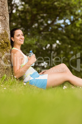 Cheerful fit brunette sitting against a tree holding a bottle