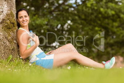 Fit brunette sitting against a tree holding her bottle