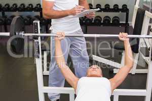 Trainer with clipboard besides woman lifting barbell in gym