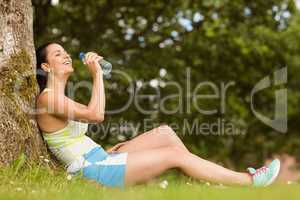 Fit brunette sitting against tree drinking water