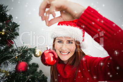 Composite image of festive redhead hanging bauble on tree