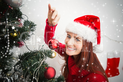 Composite image of festive redhead hanging bauble on tree