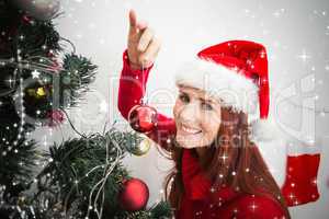 Composite image of festive redhead hanging bauble on tree