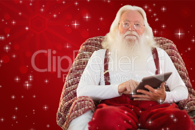 Composite image of smiling santa using tablet on the armchair