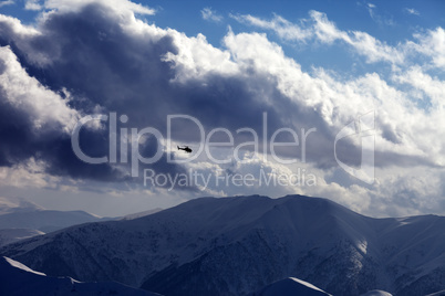 Helicopter in cloudy sky and winter mountains in evening