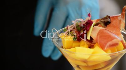 Hands of professional chef decorating cup mango fruit and ham with herbs