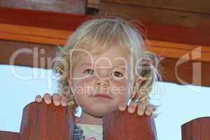Portrait of cute blond girl holding on to wooden fence.