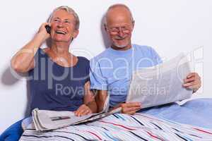 Senior Couple with Tablet and Newspaper in Bed