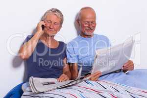 Senior Couple with Tablet and Newspaper in Bed