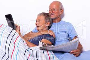 Senior Couple with Tablet and Newspaper in Bed