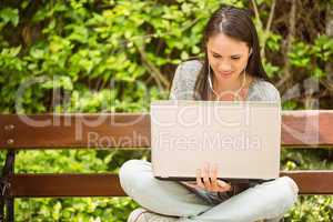 Smiling student sitting on bench listening music and using lapto