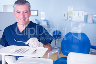 Dentist in blue scrubs smiling at camera holding tools