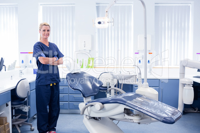 Dentist in blue scrubs standing with arms crossed beside chair