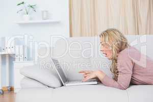 Surprised blonde lying on couch using laptop