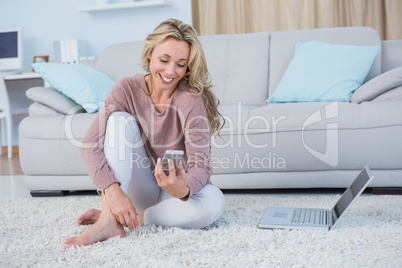 Smiling blonde sitting on carpet using smartphone