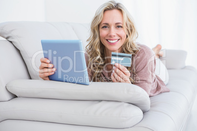 Smiling blonde lying on couch shopping online with tablet