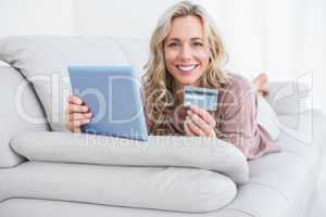 Smiling blonde lying on couch shopping online with tablet