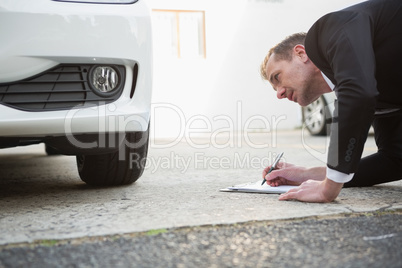 Businessman examining car tire while writing on clipboard
