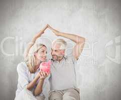 Composite image of happy couple sitting and sheltering piggy ban