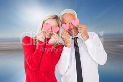 Composite image of silly couple holding hearts over their eyes