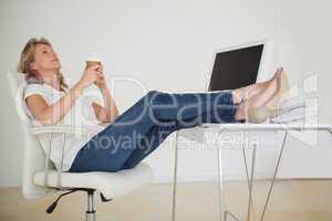 Casual businesswoman having a coffee with her feet up at desk