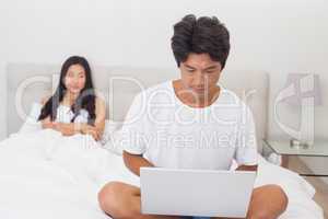Woman watching boyfriend using laptop on end of bed