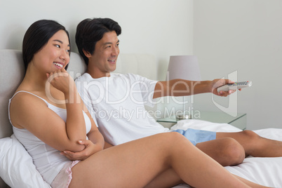 Smiling asian couple lying on bed watching tv