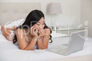 Pretty asian lying on bed talking on phone looking at laptop