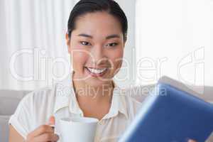 Smiling woman sitting on couch using tablet pc having coffee