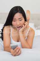 Shocked woman lying on bed holding smartphone