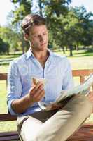Handsome man sitting on park bench eating sandwich and reading p