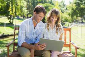 Cute couple sitting on park bench together looking at laptop