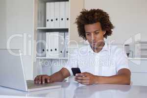 Casual businessman using his smartphone and laptop at his desk