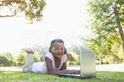 Little girl lying on grass using laptop smiling at camera