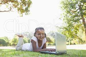 Little girl lying on grass using laptop smiling at camera