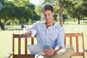 Smiling man sitting on park bench using tablet drinking coffee