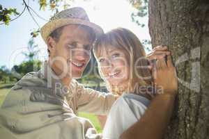 Cute couple leaning against tree in the park smiling at camera