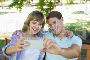 Smiling couple sitting on bench in the park taking a selfie