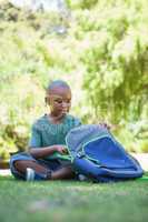 Happy schoolboy opening his schoolbag sitting on grass