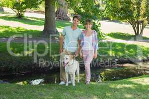 Happy couple standing with their labrador in the park smiling at