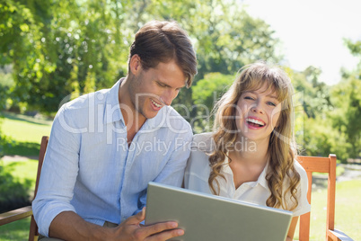 Cute couple sitting on park bench together using laptop and laug