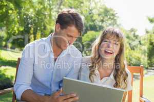 Cute couple sitting on park bench together using laptop and laug